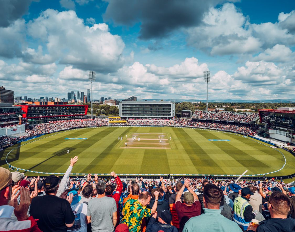 a cricket ground with people sat in the stands on a sunny day